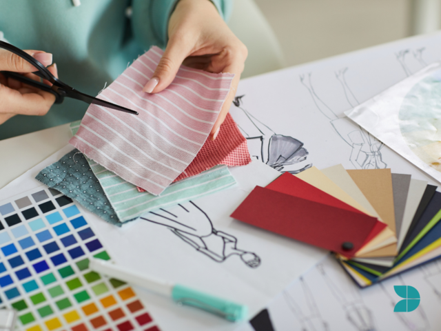 A person at a table with fashion sketches, fabric samples and color palettes using scissors to cut a piece of fabric